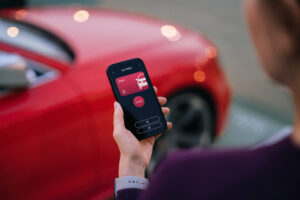 Over the shoulder view of young Asian woman using mobile app device on smartphone to unlock the doors of her intelligence car in car park. Wireless and modern technology. Smart car with digital car key concept